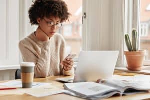 Serious female worker sits at desktop with laptop computer, surrounded with papers and journals, types sms message on smart phone device, connected to wireless internet, poses near window indoor